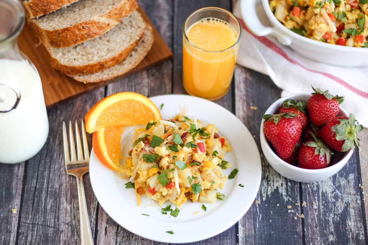 Breakfast scene with plated scramble recipe as part of whole meal with oranges, small bowl of strawberries, glass jar of milk, glass of OJ, sliced wheat bread, and serving dish of remaining skillet breakfast,
