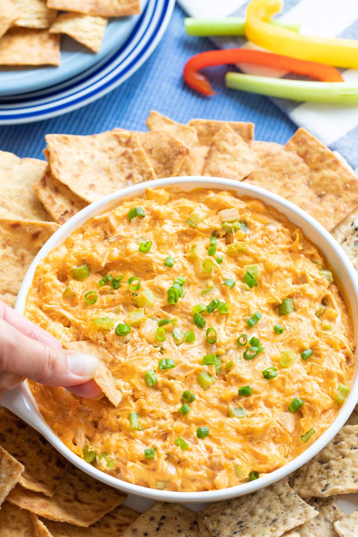 Fingers dunking a chip into the bowl of Healthy Buffalo Chicken Dip, with other chips arranged all around and vegetable stick dippers in background.