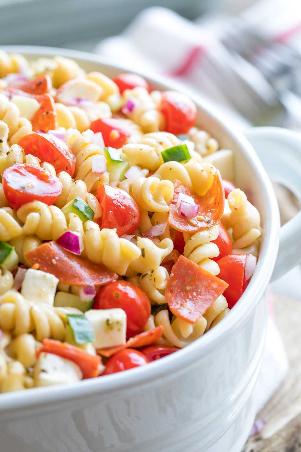 Closeup of the right side of a white serving bowl full of this pasta salad with forks and a red-striped cloth in the background..