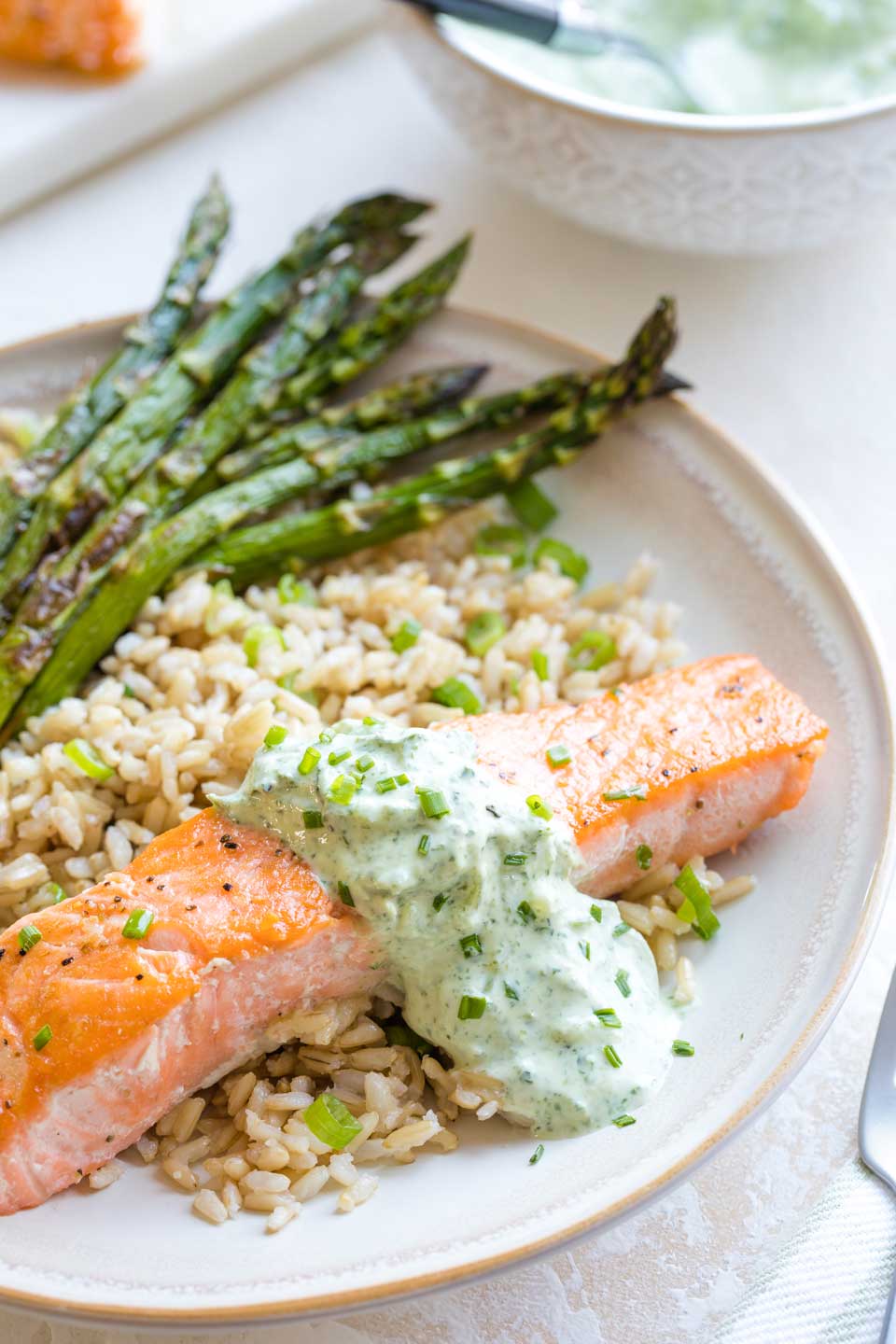 Closeup of the finished recipe, with the pan seared salmon arranged on a rimmed dinner plate, served with rice and asparagus, with utensils and additinal sauce just visible at the edges of the photo.