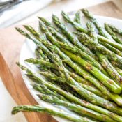 Partial view of the top portion of the roasted asparagus spears after they're out of the oven and pile on a white serving platter that's sitting on a wooden cutting board.